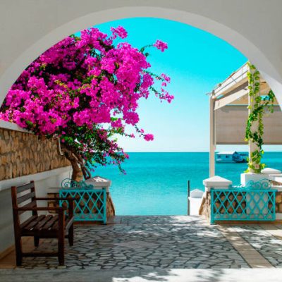 A view of the blue sea of ​​Tunisia through a white archway with pink blooming flowers in the foreground. In the distance a house on the water.